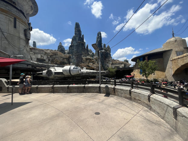 Das Millennium Falken bei Star Wars: Galaxy's Edge mit Besuchern, Schirmen, Zaun, einer Straßenlaterne, einem Schild, Bäumen, Hügeln, Drähten und einer bewölkten Himmel.
