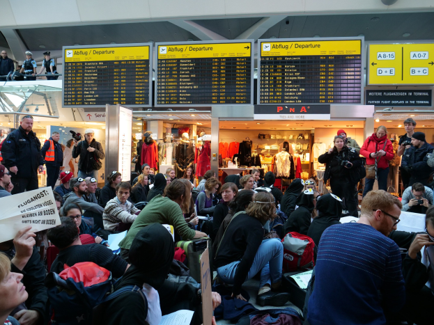 Menschen sitzen und stehen in einem Flughafen während einer Demonstration, mit Informationsschildern, Schaufensterpuppen in Kleidern und Deckenleuchten im Hintergrund.
