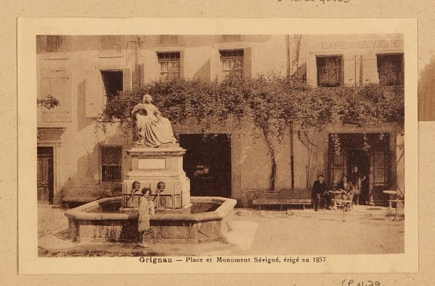 Ein altes Foto eines Brunnens vor einem Gebäude, mit einer Statue auf einem Sockel in der Mitte, Menschen auf Bänken um den Brunnen herum und einem Baum im Hintergrund, mit Text unten auf dem Bild.