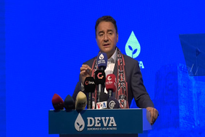 Ein Mann in einem Mantel und einer Mütze steht an einem Podium mit Mikrofonen, ein Schild mit der Aufschrift "deva-Geschäftsführer sagt, es sei keine gute Idee für die Zukunft", ein Objekt und ein Bildschirm im Hintergrund.