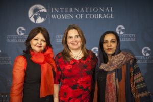 Drei Frauen stehen zusammen und lächeln vor einem Banner mit der Aufschrift "International Women of Courage".