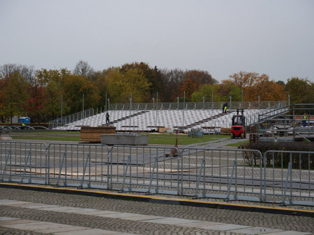 Großes Stadion mit Metallgeländern, umgeben von Bäumen unter einem klaren blauen Himmel, mit einigen Menschen und Fahrzeugen im Vordergrund auf saftig grünem Rasen.