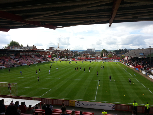 Ein Fußballspiel im St. Mary's Stadium, mit Zuschauern auf den Tribünen und in der Nähe des Torpfostens, umgeben von Gebäuden, Bäumen und einem bewölkten Himmel.