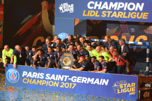 Spieler von Paris Saint-Germain feiern mit der Ligue-1-Trophäe, tragen Medaillen, stehen vor einer Tafel mit Text und Logos, mit einem Netz auf der rechten Seite und einem Bildschirm im Hintergrund.