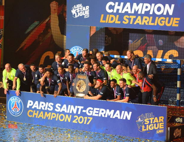 Spieler von Paris Saint-Germain feiern mit der Ligue-1-Trophäe, tragen Medaillen, stehen vor einer Tafel mit Text und Logos, mit einem Netz auf der rechten Seite und einem Bildschirm im Hintergrund.