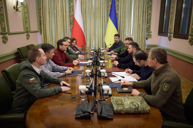 Eine Gruppe von Männern in einem formellen Setting, die um einen Holztisch mit Mikrofonen und Dokumenten sitzen und diskutieren, mit Flaggen und Beleuchtung im Hintergrund.