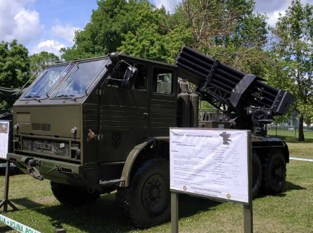 Ein Militärlastwagen mit einer Rakete darauf steht auf einer Wiese umgeben von Bäumen unter einem klaren blauen Himmel, mit einer Texttafel davor, wahrscheinlich Teil einer russischen Militärausstellung.