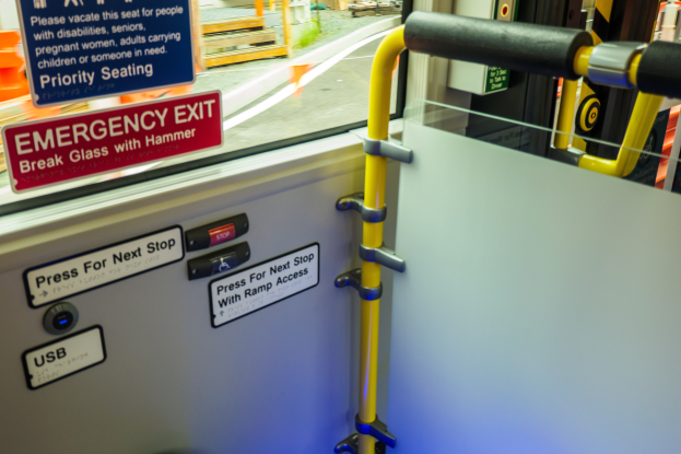 Notausgangsschild an der Seite eines Zuges, mit Türgriff im Vordergrund und einem Glasfenster, das Straße, Pfosten und andere Gegenstände draußen zeigt.