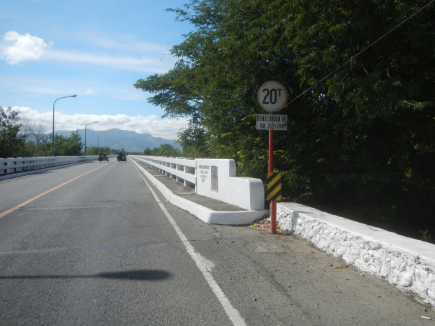 Eine mehrspurige Straße mit fahrenden Fahrzeugen, ein Tempolimit-Schild, Metallgeländer, hohe Laternen, Bäume und ein bewölkter Himmel im Hintergrund.