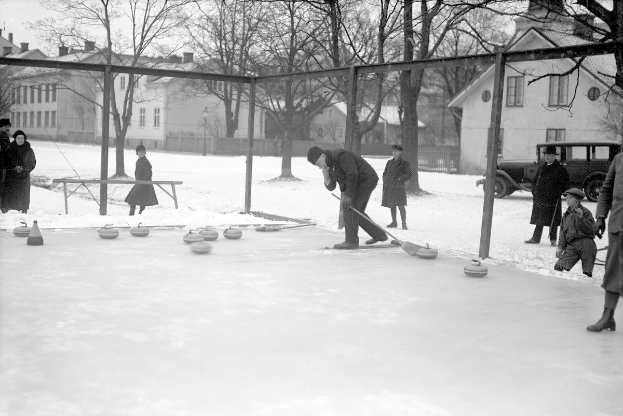 Schwarzes Bild von Menschen, die Curling auf einer Eisbahn spielen, umgeben von einem Zaun, mit einer nahen Bank, Bäumen, Gebäuden mit Fenstern und einem geparkten Fahrzeug im Hintergrund.