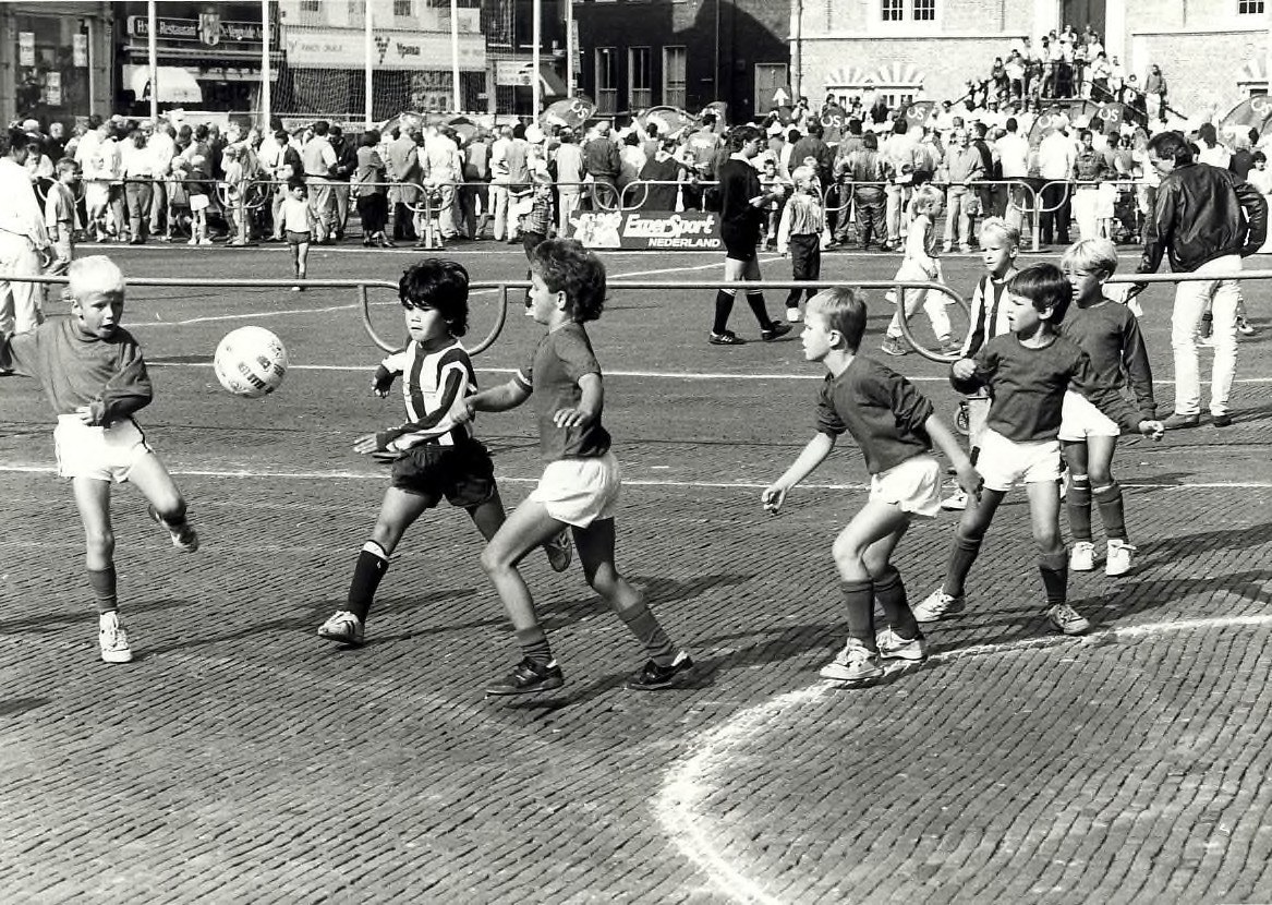 Schwarzes Foto von jungen Jungs, die auf der Straße Fußball spielen, mit Zuschauern hinter einem Geländer, Gebäuden und Texttafeln im Hintergrund.