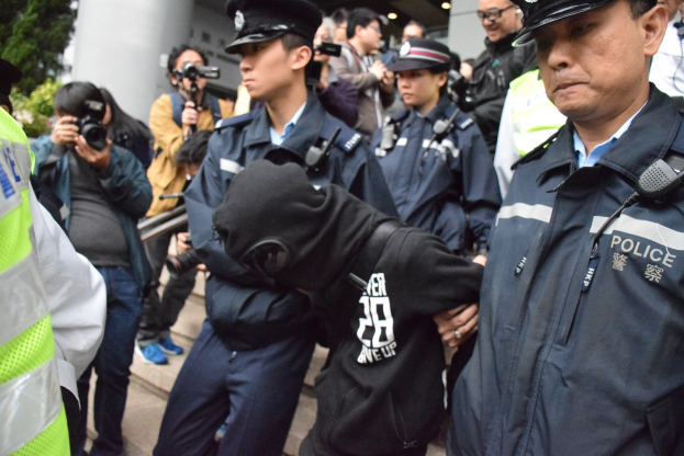 Eine Gruppe von Polizeibeamten steht vor einer Menge vor einem Gebäude mit Säulen und Deckenbeleuchtung, mit Bäumen links sichtbar.