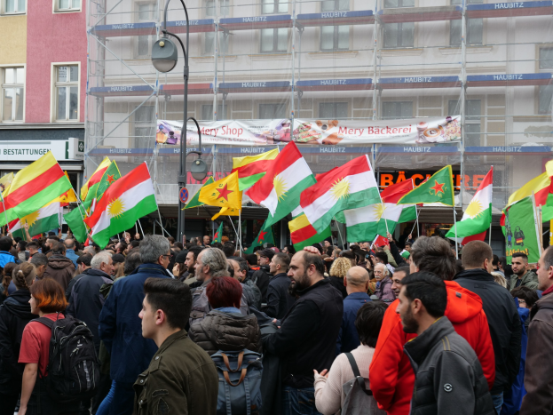 Große Gruppe von Menschen vor einem Gebäude mit einigen, die kurdische Flaggen halten, vorne ein Laternenmast und ein Banner mit Text im Hintergrund.