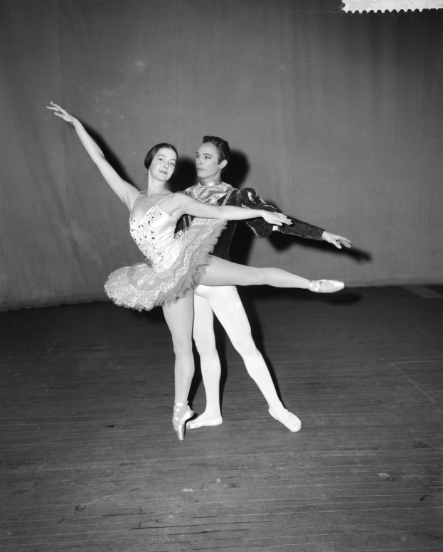 Schwarzes und weißes Foto eines Mannes in einem weißen Hemd und schwarzen Hosen, der mit einer Frau in einem unbekannten Outfit auf einer Bühne tanzt, deren Körper in perfekter Harmonie synchronisiert sind.