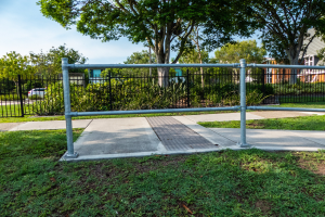 Ein Metallzaun in einer grasbewachsenen Fläche, umgeben von Pflanzen, Bäumen und einem Auto auf der Straße, mit Gebäuden und einem klaren blauen Himmel im Hintergrund, wahrscheinlich Teil eines Spielplatzes.