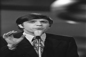 Ein Mann in einem schwarzen Anzug, weißem Hemd und gemustertem Schlips spricht in ein Mikrofon, mit einem ernsten Gesichtsausdruck und zurückgekämmtem Haar, vor einem unscharfen Hintergrund.