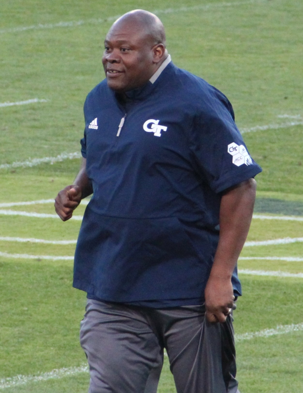 Ein Mann in einem blauen Hemd und grauen Hosen steht auf einem grünen Feld, lächelt mit einem selbstbewussten und entschlossenen Ausdruck und sieht aus wie ein Fußballtrainer.