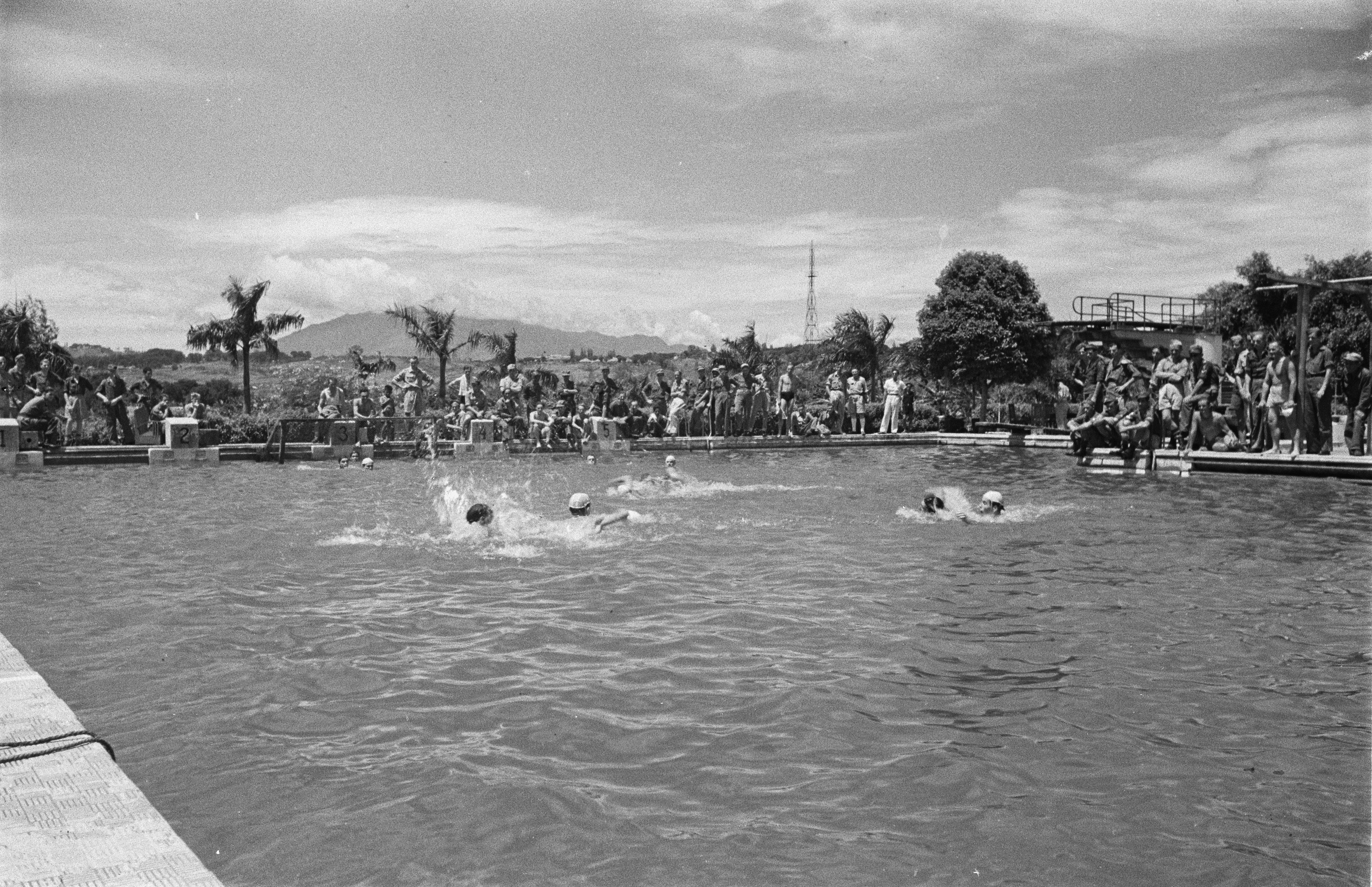 Schwarzes und weißes Foto von Menschen, die in einem Pool schwimmen, mit Zuschauern, einem Zaun, Pfählen, einem Turm, Bäumen, Hügeln und einem bewölkten Himmel.