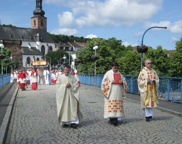 Gruppe von Priestern, die eine Straße mit Laternenmasten, Geländern und Bäumen entlanggehen, im Hintergrund Gebäude und einen bewölkten Himmel; einer der Priester hält ein Buch.