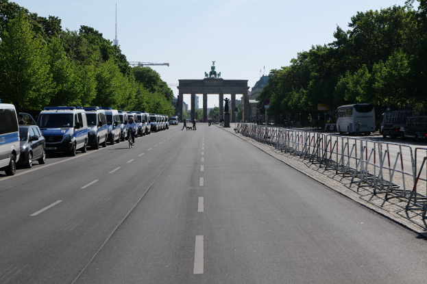 Lange Reihe von Polizeiwagen am Straßenrand vor dem Brandenburger Tor in Berlin geparkt, mit Menschen auf Fahrrädern und in der Nähe stehend, Absperrungen, Bäume, ein Bogen mit Statuen im Hintergrund und sichtbarem Himmel.