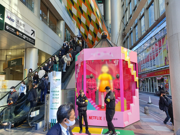 Eine Gruppe von Menschen steht vor einem Gebäude mit Glaswänden, Säulen und Texttafeln, einige tragen Masken, in der Nähe einer Rolltreppe mit Menschen darauf; ein pinker Kasten in der Mitte zeigt Schaufensterpuppen und trägt die Aufschrift "Netflix's new Netflix booth in Tokyo."