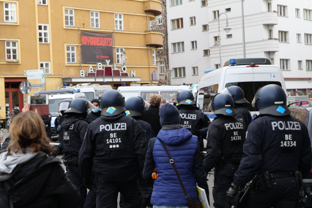 Polizeibeamte in Uniform vor einer Menge von Demonstranten mit Kameras, mit Fahrzeugen, Gebäuden und einem Banner im Hintergrund während einer Demonstration in Berlin, Deutschland.