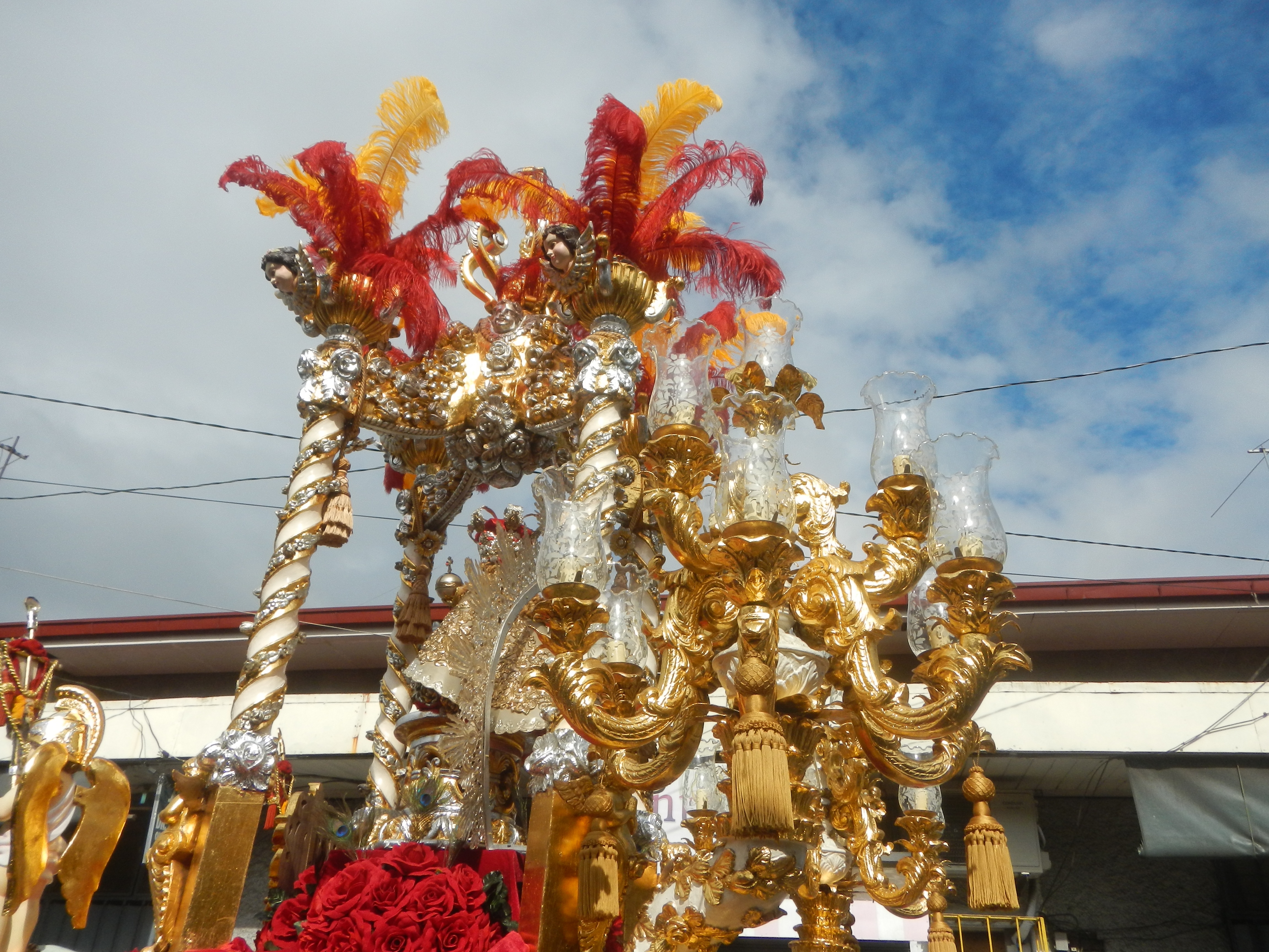 Ein großes goldenes und rotes Schwimmwagen, geschmückt mit Blumen und anderen Dekorationen, in einem Karnevalsumzug, mit einem Gebäude, Strommasten mit Drähten und einem bewölkten Himmel im Hintergrund.