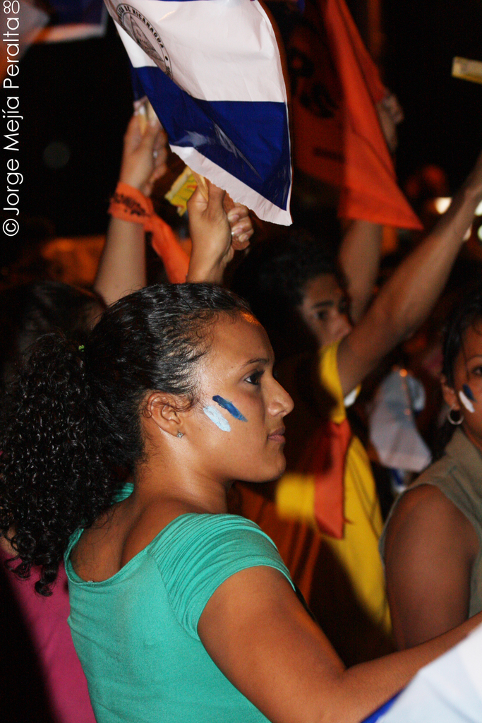 Eine Frau in einem grünen T-Shirt steht bei einer Protestaktion und hält ein weißes Schild in der Hand, während viele andere Menschen hinter ihr stehen.