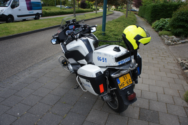 Ein polizeiliches Motorrad am Straßenrand mit einem Helm darauf, umgeben von Bäumen, Polen, Gebäuden, Pflanzen, Gras, Steinen und anderen Fahrzeugen.