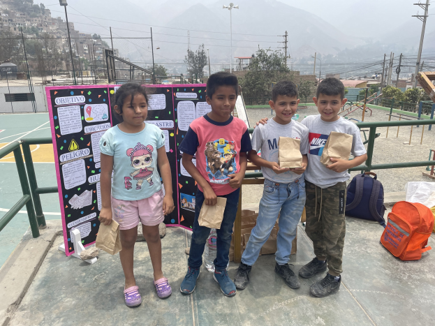 Gruppe von Kindern auf einem Basketballfeld mit Papiertüten, Tüten und Gegenstände auf der rechten Seite und eine Tafel mit Text auf der linken Seite, vor einem Hintergrund aus Zäunen, Pfosten, Straßenlaternen, Bäumen, Gebäuden, Hügeln und Himmel.