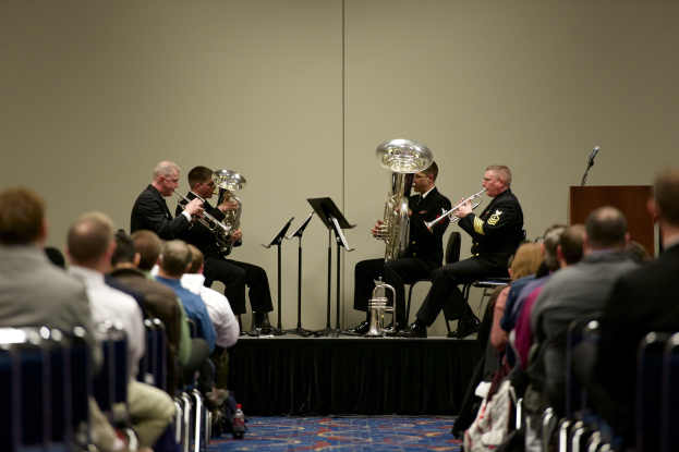 Gruppe von Männern, die auf einer Bühne Blasinstrumente spielen, mit Notenständern, einem Podium mit Mikrofon und einem Publikum, das auf Stühlen sitzt.