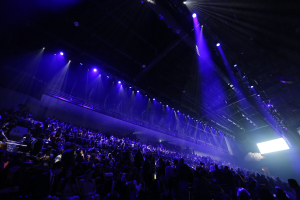 Eine große Menschenmenge in einem Konzertsaal, die einer Bühne mit Deckenleuchten und einem Bildschirm auf der rechten Seite gegenüber sitzt.