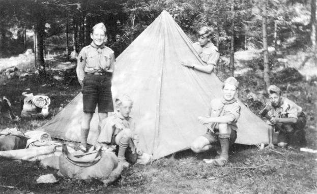 Schwarz-weißes Bild von Pfadfindern in Uniformen, die sich um ein Zelt in einem bewaldeten Gebiet versammelt haben, einige sitzen auf dem Boden, während andere stehen, mit verstreuten Taschen und Gegenständen in der Nähe.
