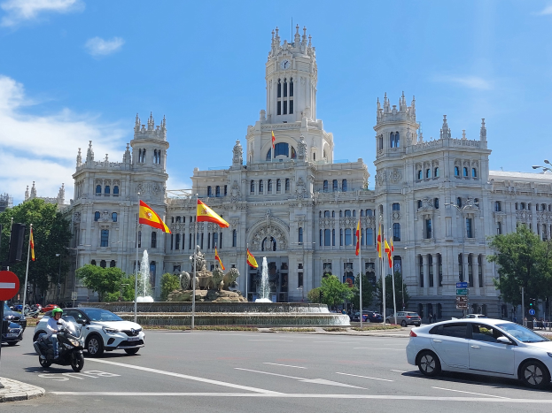 Eine belebte Straße in Madrid, Spanien, mit Fahrzeugen, einem Radfahrer, einer Schautafel, Mästängen, Lichtern, Bäumen, Flaggen, einem Brunnen und einem großen Gebäude mit einem Uhrenturm unter einem bewölktem Himmel.