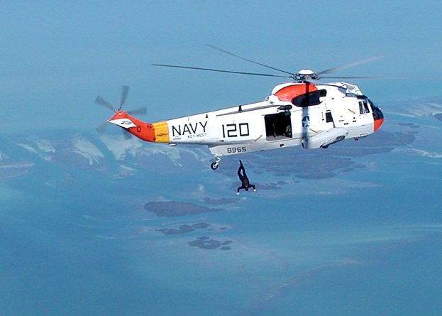 Hubschrauber fliegt über Wasser mit einer Person darunter und Himmel im Hintergrund, beschriftet als Rettungshubschrauber.