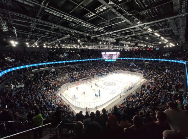 Eishockeyspiel in einer großen Arena mit Zuschauern in den Rängen, Geländern, Überbildschirm und heller Beleuchtung, identifiziert als die O2 Arena in London.