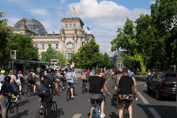 Eine Gruppe von Menschen, die auf Fahrrädern eine von Bäumen gesäumte Straße in Berlin, Deutschland, entlangfahren, mit einer Bushaltestelle auf der rechten Seite und einer Flagge, die auf einem nahegelegenen Gebäude weht.