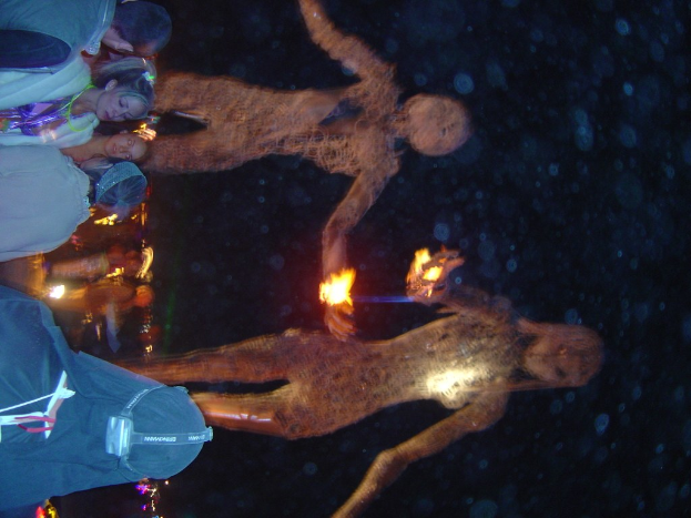 Menschen vor brennenden Statuen mit Lichtern im Hintergrund.