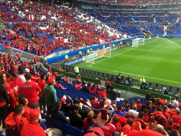 Eine große Menschenmenge in einem Stadion schaut einem Fußballspiel zu, mit Sitzplätzen und Stehplätzen, Bannern, einem Torpfosten und Deckenbeleuchtung unter einer elektrisierenden Atmosphäre.