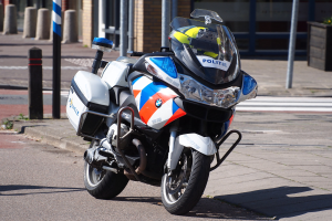 Ein polizeiliches Motorrad am Straßenrand mit einem Helm darauf, mit Masten, Pflanzen und einem Gebäude im Hintergrund.