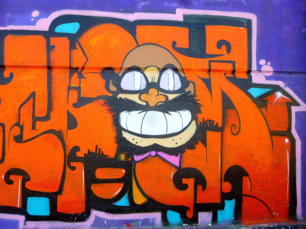 Graffiti-Gemälde eines Cartoon-Charakters mit großen Augen, einem breiten Grinsen und einem verschmitzten Ausdruck an der Seite eines Gebäudes, mit leuchtenden Farben und einer mutigen Begleittext.