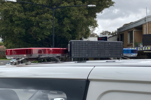 Ein Polizeiwagen mit eingeschaltetem Blaulicht steht am Straßenrand, umgeben von Gebäuden, Bäumen und Masten, mit Texttafeln in der Nähe, unter einem bewölkten Himmel, wahrscheinlich nach einem tödlichen Schulschießen in Brisbane.