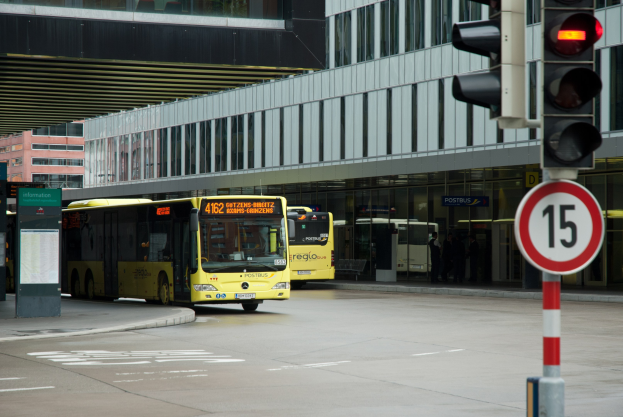 Zwei gelbe Busse fahren auf einer Straße mit hohen Gebäuden, einer Ampel auf der rechten Seite und Fußgängern auf dem Gehweg.