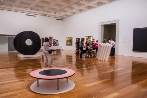 Eine Gruppe von Menschen in einer Kunstgalerie mit einer Person im Rollstuhl in der Mitte, umgeben von gerahmten Fotos an den Wänden unter beleuchteter Decke, mit einem schwarzen Installationsobjekt links.