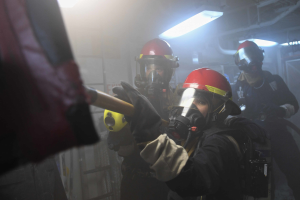 Eine Gruppe von Menschen in Gasmasken und Helmen, einer hält ein Objekt, steht in einer gefährlichen Umgebung mit Lampen und Rohren im Hintergrund.