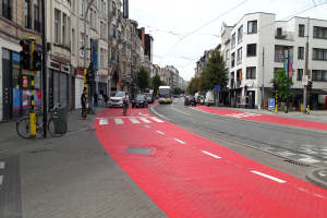 Eine Stadtstraße mit einem roten Fahrradweg, Fahrzeuge auf der Straße, Fußgänger auf dem Gehweg, Gebäude mit Fenstern auf beiden Seiten, Bäume und einen klaren blauen Himmel im Hintergrund.