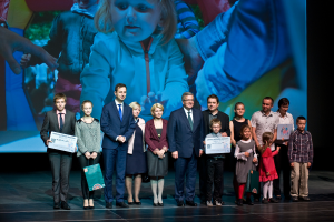 Eine Gruppe von Menschen auf einer Bühne, einige halten Taschen und Urkunden, im Hintergrund ein Bildschirm mit einem Bild von einem Kind und anderen Menschen.