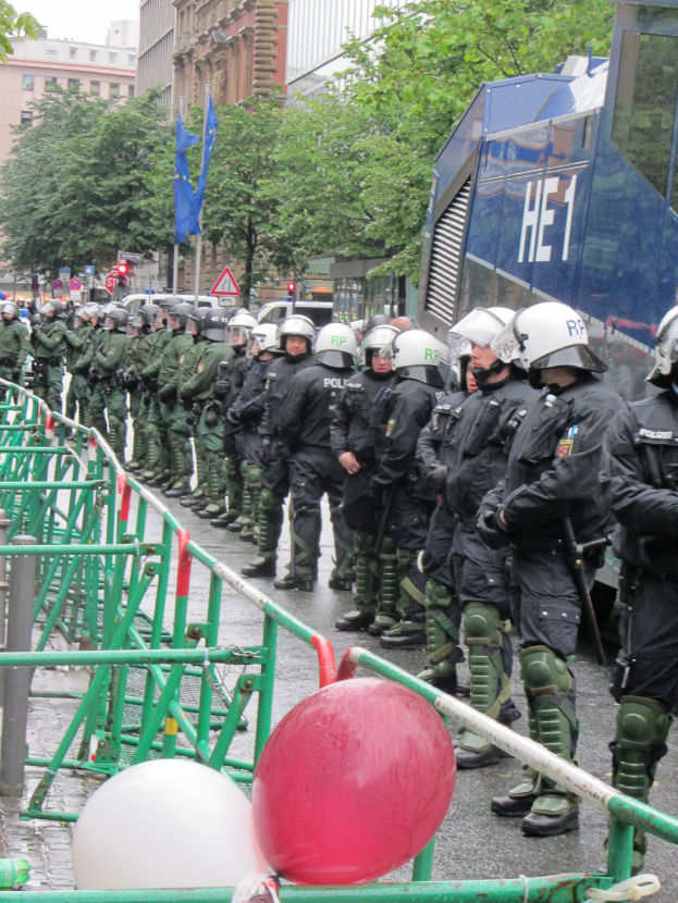 Polizeibeamte in Helmen und Uniformen stehen hinter einer Barriere mit Waffen, umgeben von Geländern, Luftballons, Bäumen, Gebäuden, Flaggen und einem klaren Himmel.
