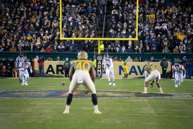 Fußballspieler in Helmen und Uniformen auf einem Feld mit Zuschauern in den Rängen und Stadionwerbung im Hintergrund.