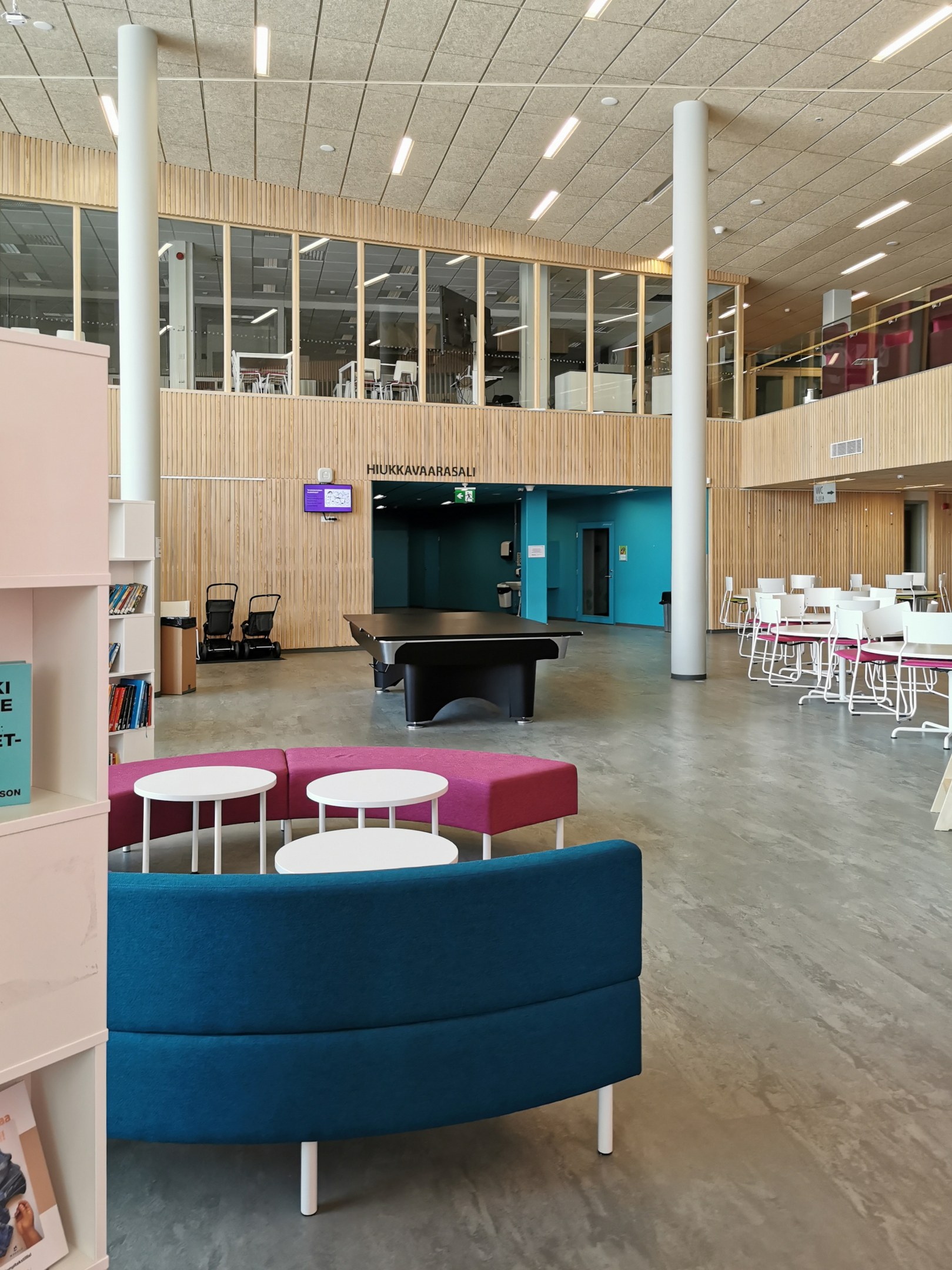 Ein geräumiger Schulinnenraum mit Tischen, Stühlen, Sofas, Hockern, Bücherregalen, Säulen, Deckenleuchten und einem Wandfernseher, der an eine Bibliothek erinnert.
